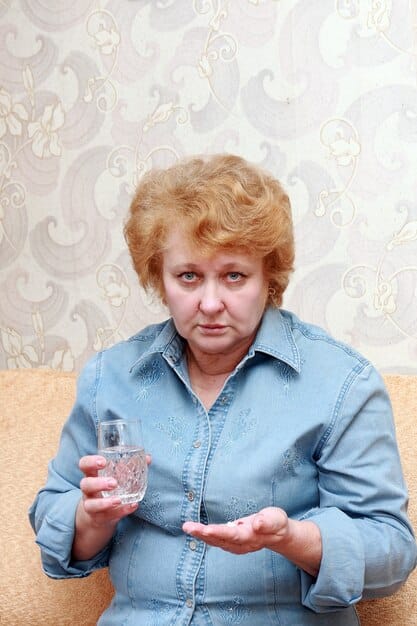 An elderly woman sitting indoors, sipping a glass of water, with a concerned expression. A fan points towards her, and the room is dimly lit. The scene conveys the vulnerability of older people during heat waves.