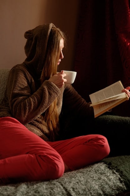 A person reading a physical book in a comfortable armchair, with a warm cup of tea on a side table. Sunlight streams through the window, creating a peaceful and inviting reading nook.