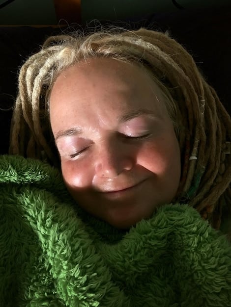 An eye-level close-up shot of a person's relaxed face in a sunlit environment, with their eyes closed and a gentle smile subtly showing on their face, indicating peacefulness and contentment. The soft lighting accentuates their serene expression.