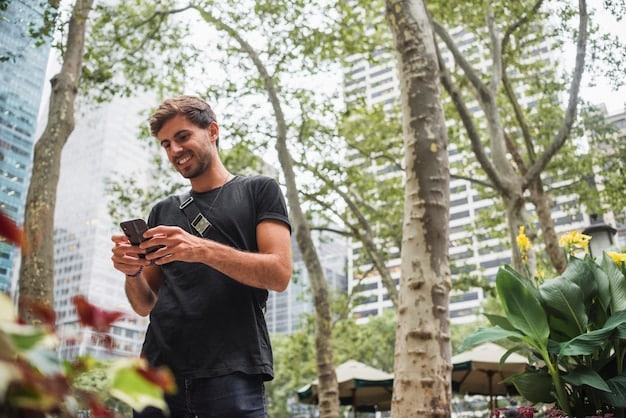 A young adult smiling and looking away from their phone, standing in a sunny park with trees and other people in the background, suggesting a healthy outdoor activity.